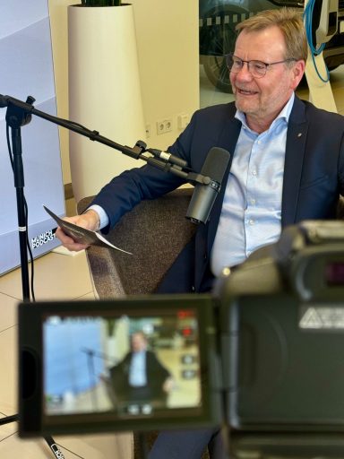 Mann sitzt in einem Stuhl, spricht in ein Mikrofon und hält ein Tablet in der Hand.