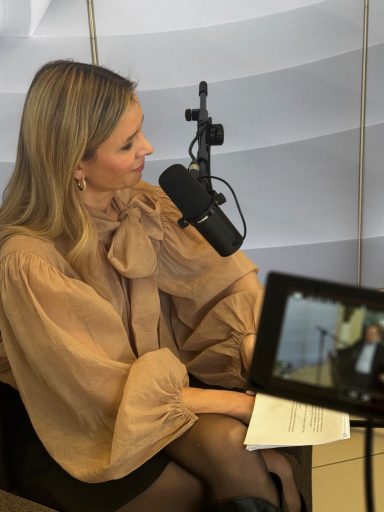 Frau mit blonder Haarfarbe trägt eine helle Bluse und spricht in ein Mikrofon.