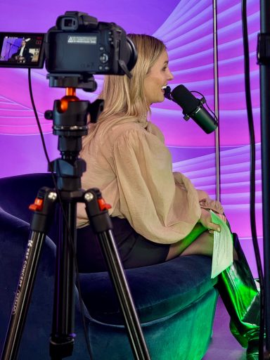 Frau mit Mikrofon sitzt auf einem grünen Sessel vor Kamera und lila Hintergrund.