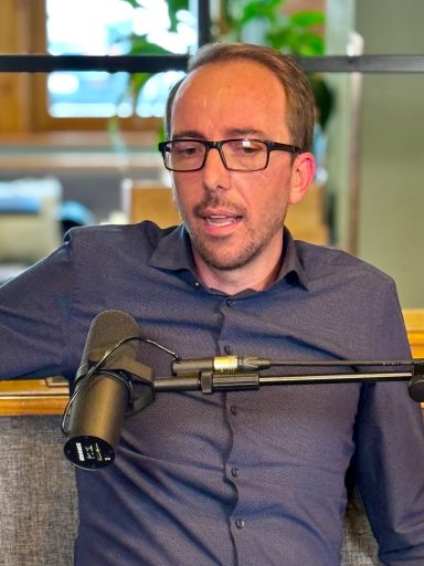 Mann mit Brille sitzt lächelnd neben einem Mikrofon in einem modernen Raum.