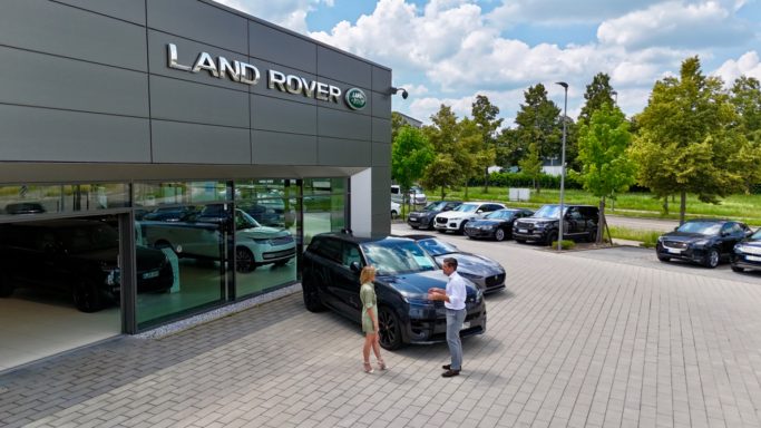 Mann und Frau sprechen vor einem modernen Autohaus mit mehreren Fahrzeugen.