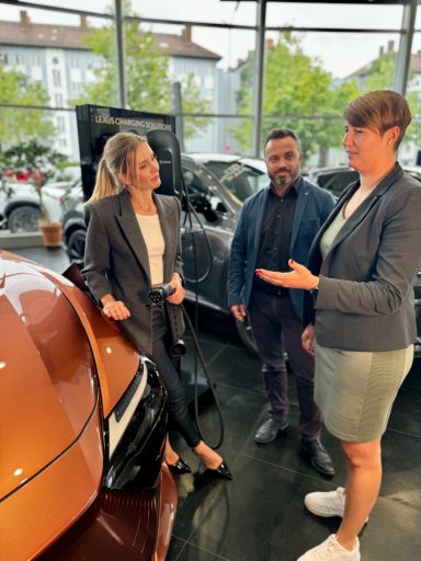 Drei Personen im Gespräch in einem modernen Autohaus, vor einem orangefarbenen Auto.