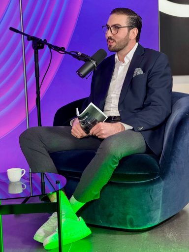 Mann in Anzug mit Mikrofon, sitzt auf blauer Couch und hält ein Buch in der Hand.